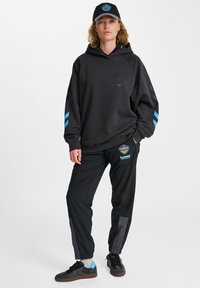 Svart huvtröja med blå detaljer, prydd med en logotyp. Svarta sweatpants med branding och en grå panel. Mörka skor med blå accenter.