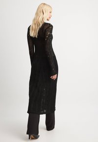Black, long-sleeve knit dress with an open, textured design and vertical lines. Worn over black pants and heeled shoes.