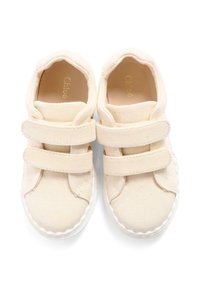 Zapatillas de lona beige con dos tiras de velcro, punta redonda y suelas de goma blancas. Textura suave con un patrón salpicado sutil. En el interior presenta un logo dorado.
