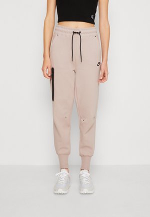 Persoon die beige Nike joggers draagt met een zwart trekkoord, zwart crop top en witte sneakers, staand tegen een effen witte achtergrond.