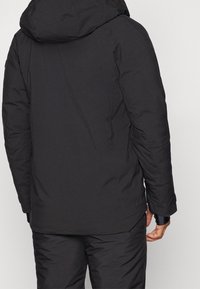 Osoba v černé zateplené bundě s kapucí a odpovídajících černých kalhotách, zobrazená zezadu na jednobarevném pozadí.