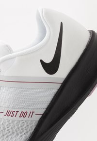 Hvid atletiksko med en struktureret mesh-overdel, sort Nike swoosh-logo, "JUST DO IT" tekst i rød og en sort gummisål.