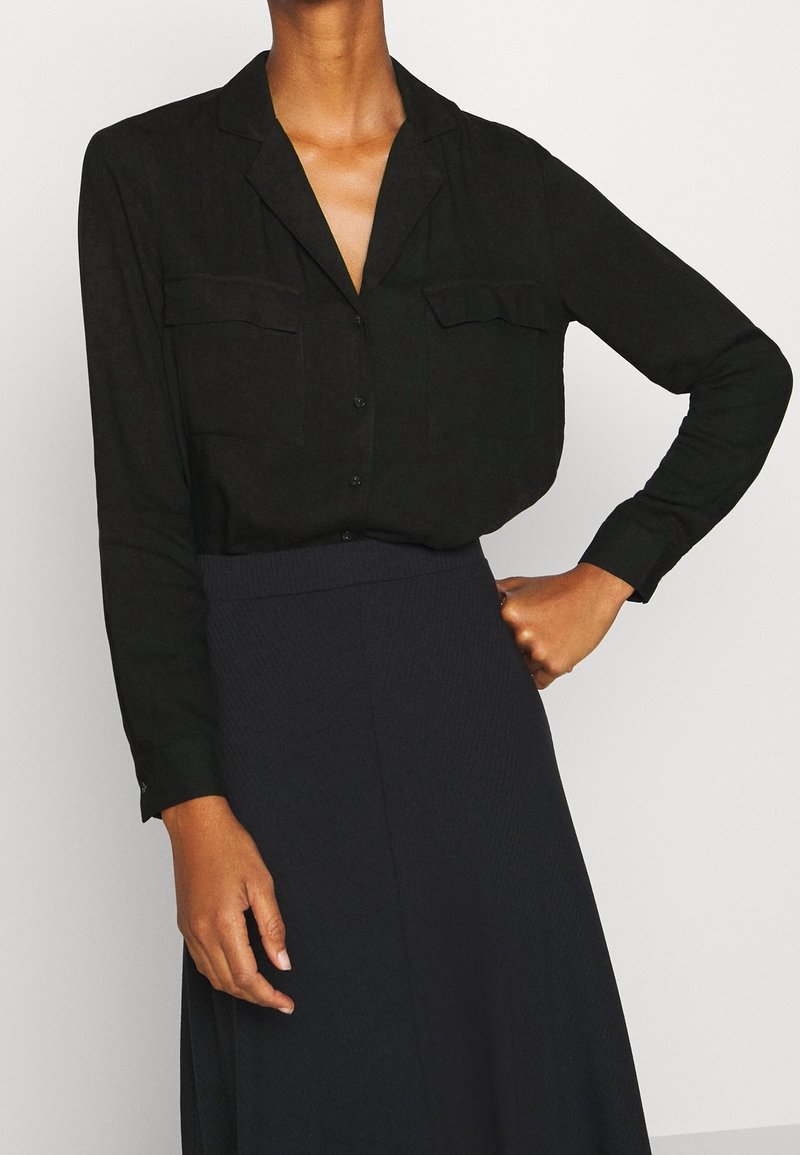 Femme portant un chemisier noir boutonné avec des poches poitrine et une jupe sombre taille haute, posant avec une main sur la hanche.
