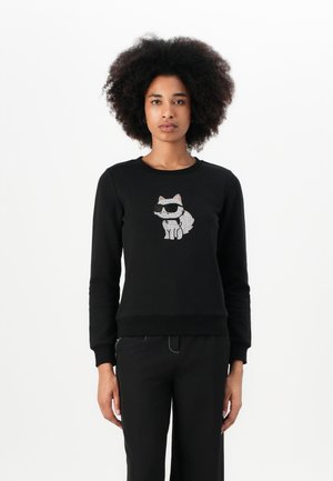 Felpa nera in cotone, con una grafica di un gatto in occhiali da sole. Collo a giro e polsini e orlo a costine. Indossata con pantaloni neri.