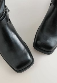 Mustad muste kingad, mis on valmistatud siledast kunstnahast, ruuduotsaga kujundusega, kontrastse õmblusega ja dekoratiivsete külgbuksidega.