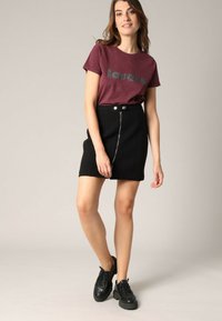 T-shirt bordeaux avec "lovers" imprimé dans un design brillant, accompagné d'une mini-jupe noire avec fermeture éclair et bouton à l'avant. Baskets noires.