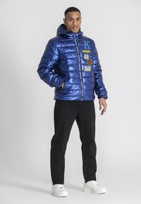 Blå pufferjacka med glänsande textur, med flera lappar och huva. Kombinerad med svarta byxor och vita sneakers.