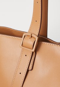 Tan leren handtas met een dikke schouderriem en rechthoekige gespdetails, voorzien van nauwkeurige stiksels en een gladde, matte afwerking.
