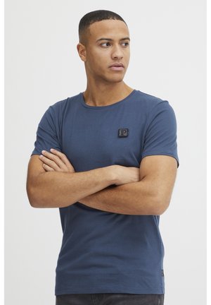 Camiseta de color azul marino con cuello redondo y mangas cortas. Presenta un pequeño parche de logo negro en el pecho. Tejido suave y liso.