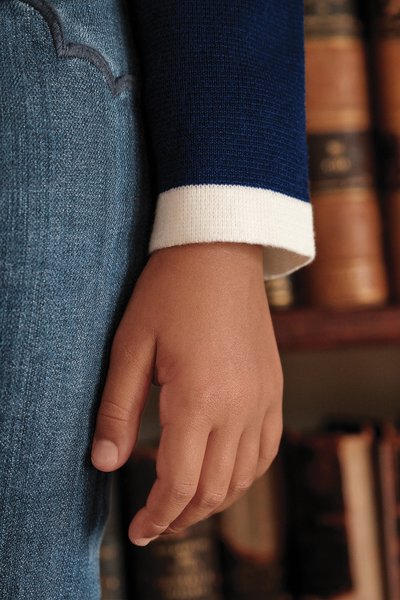 Pullover de malha azul-marinha com punhos brancos, combinado com jeans azul. Uma mão repousa sobre o tecido texturizado dos jeans. Livros ao fundo.