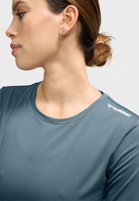 Teal sportshirt met een ronde hals en een gestructureerde stof. De linkerschouder heeft het witte logo van "hummel." Oor met zichtbaar sieraad.