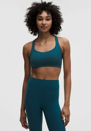 Donna con capelli ricci che indossa un reggiseno sportivo turchese e leggings a vita alta, in piedi davanti a uno sfondo chiaro e semplice.