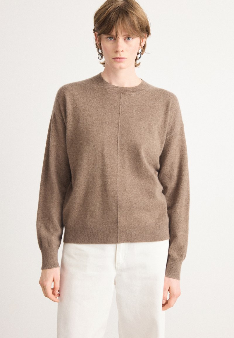 Καφέ πλέκτο πουλόβερ με στρογγυλή λαιμόκοψη, χαμηλούς ώμους και κεντρική ραφή. Φορεμένο με ανοιχτόχρωμο παντελόνι. Μαλακή υφή.