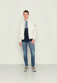 Casaco bomber branco, t-shirt azul-marinho com logótipo, jeans azuis desgastados, sapatilhas castanhas com detalhes brancos. Modelo de pé contra um fundo neutro.