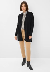 Schwarzer Strickcardigan mit offenem Frontbereich, geripptem Kragen und langen Ärmeln, getragen über einem gestreiften Hemd und beigen Hosen, kombiniert mit schwarzen Stiefeletten.