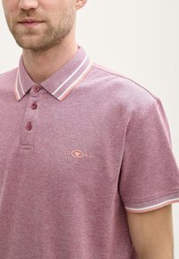 Polo shirt i en meleret burgundy farve med en to-knap lukning, ribbet krave med pink og hvide striber samt broderet logo-detalje.
