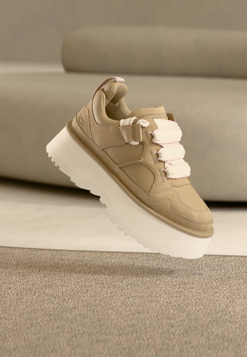 Μπεζ αθλητικό παπούτσι με χοντρή λευκή σόλα, ανώτερο δέρμα με υφή,  υφασμάτινα κορδόνια και θηλιά στο πίσω μέρος για εύκολη εφαρμογή. Πλέει πάνω από μια μαλακή επιφάνεια.