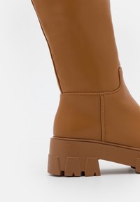 Brązowy półbut z cholewką do połowy łydki, z masywną, żłobioną podeszwą i widocznym przeszyciem, pokazany z boku na białym tle.