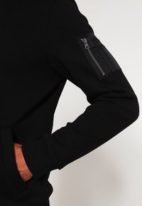 Personne portant une veste noire à manches longues avec une poche zippée sur la manche supérieure et la main dans la poche latérale.