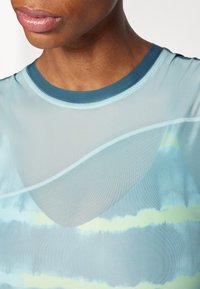 Lengvas, permatomas sportinis marškinėlis akvamarino spalvos su apvalia iškirpte, pasižymintis bangų raštais šviesiai geltonos ir mėlynos spalvos, bei kontrastingais apvadais.
