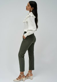 Blusa blanca con detalle de volantes, pantalones verde oliva y zapatos de tacón cuadrado beige; perfil de pie con cabello largo y oscuro y pendientes de oro.