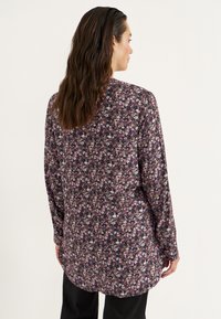 Dunkelbluse mit langen Ärmeln, die ein mehrfarbiges Blumenmuster auf schwarzem Hintergrund zeigt, mit einem leicht abgerundeten Saum und lockerer Passform.