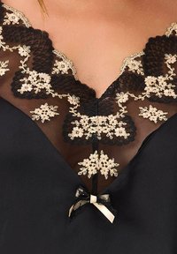 Czarna odzież z głębokim dekoltem w kształcie litery V, zdobiona misternym beżowym haftem florystycznym i koronkowym wykończeniem, z małą kokardką przy dekolcie.