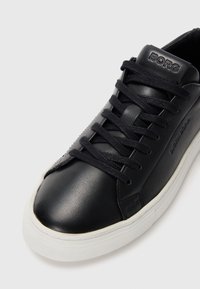 Svart lærsneaker med tonale snørebånd, gummisåle og preget logo på tungen. Jevn tekstur og rund tådesign.
