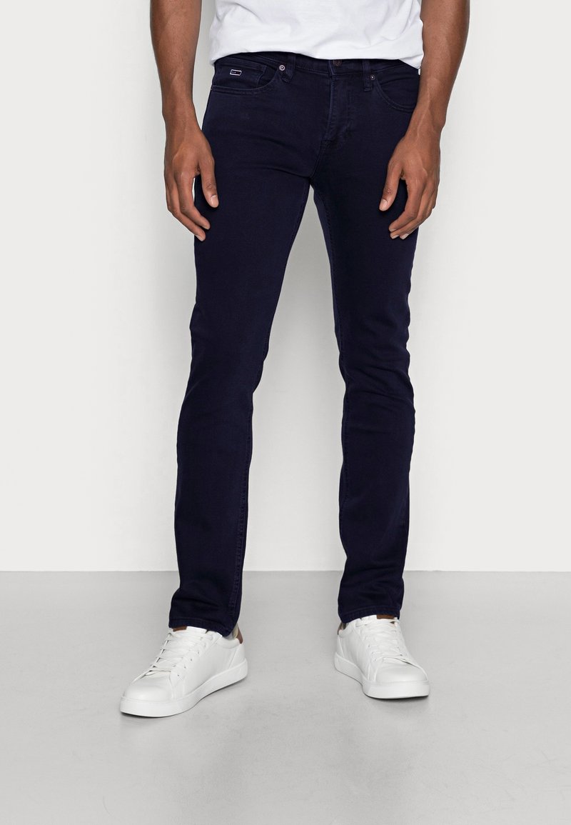 Jeans de mezclilla azul oscuro con un corte slim. Cuenta con cinco bolsillos, cierre de cremallera y detalles de costura sutiles. Combinados con zapatillas blancas.