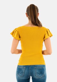 Top amarillo acanalado con mangas cortas y volantes, diseño ajustado y escote amplio. Combinado con jeans de mezclilla azul con costuras sutiles.