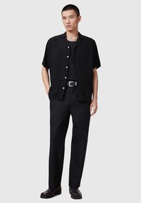 Vyras, vilkintis juodą tekstūruotą trumpą rankovę marškinėlius, juodas kelnes su diržu, juodus batus ir sidabrinius papuošalus, stovi prieš paprastą foną.