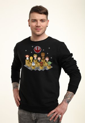 Mand iført sort sweatshirt med pixelkunstfigurer fra Star Wars arrangeret på podieblokke med Rebel Alliance-symbolet ovenover.