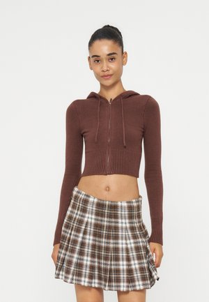 Kvinde iført en brun zip-up cropped hoodie og et brun-hvidt ternet plisseret skørt, stående mod en ensfarvet lys baggrund.