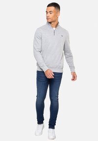 Sweatshirt gris zippé en tissu texturé. Jean slim-fit bleu marine et baskets blanches. Design simple sans motifs visibles.