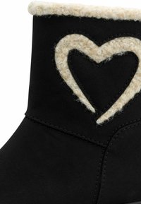 Zwarte enkellaars met een wollige crème manchet en een hartvormige uitsparing aan de zijkant, gemaakt van gestructureerd suède materiaal en contrasterende stiksels.