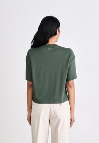 Olivengrønn bomulls T-skjorte med en cropped design, korte ermer og en liten brodert logo på baksiden. Brukt med lysebeige bukser.