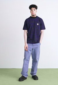 Svītrains tumši zils un purpura kokvilnas T-krekls ar īsām piedurknēm, Carhartt logo uzšuvums; apvienots ar gaiši purpura biksēm un melniem zābakiem bez šņorēm.