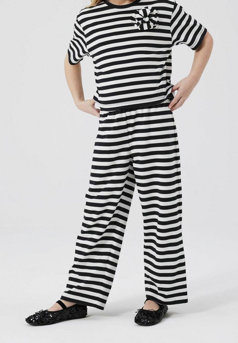 Enfant portant un haut à manches courtes rayé horizontal noir et blanc avec un pantalon large assorti et des chaussures noires à paillettes, mains sur les hanches.