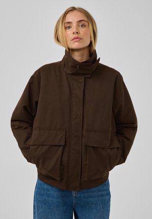 Jeune femme portant une veste marron foncé à col montant avec de grandes poches avant et un jean bleu, debout devant un fond gris uni.