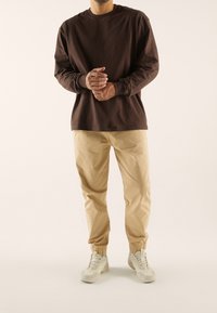 Brązowa koszula z długim rękawem, beżowe zwężane spodnie z elastycznymi mankietami oraz jasnoszare trampki. Gładka tkanina, neutralne kolory, swobodny styl.