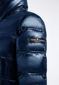 Marineblauwe pufferjack met een glanzende, gewatteerde afwerking. Heeft een capuchon, elastische manchetten en een stoffen label op de mouw. Glad van structuur.