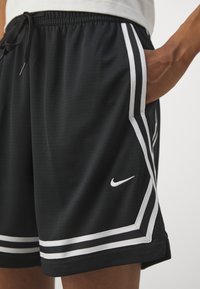 Personne portant un short de sport noir Nike avec un liseré blanc et un logo, main reposant dans la poche latérale.