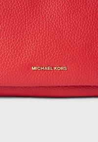 Sarkans ādas rokassomu ar teksturētu virsmu un zelta "MICHAEL KORS" logotipu priekšpusē. Kompakts dizains ar noapaļotām malām.