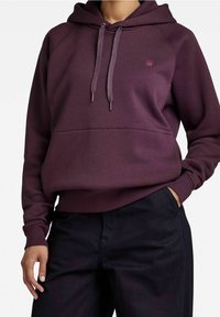 Felpa bordeaux con tasca a kangaroo, cappuccio con cordino e polsini a costine. Presenta un logo discreto sul petto. Materiale in misto cotone.