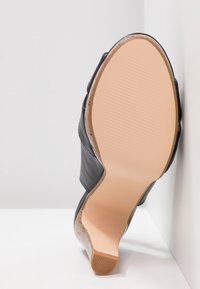 Chaussure à talon en cuir noir positionnée semelle vers le haut, montrant une semelle beige lisse et un talon bloc bas sur fond blanc.