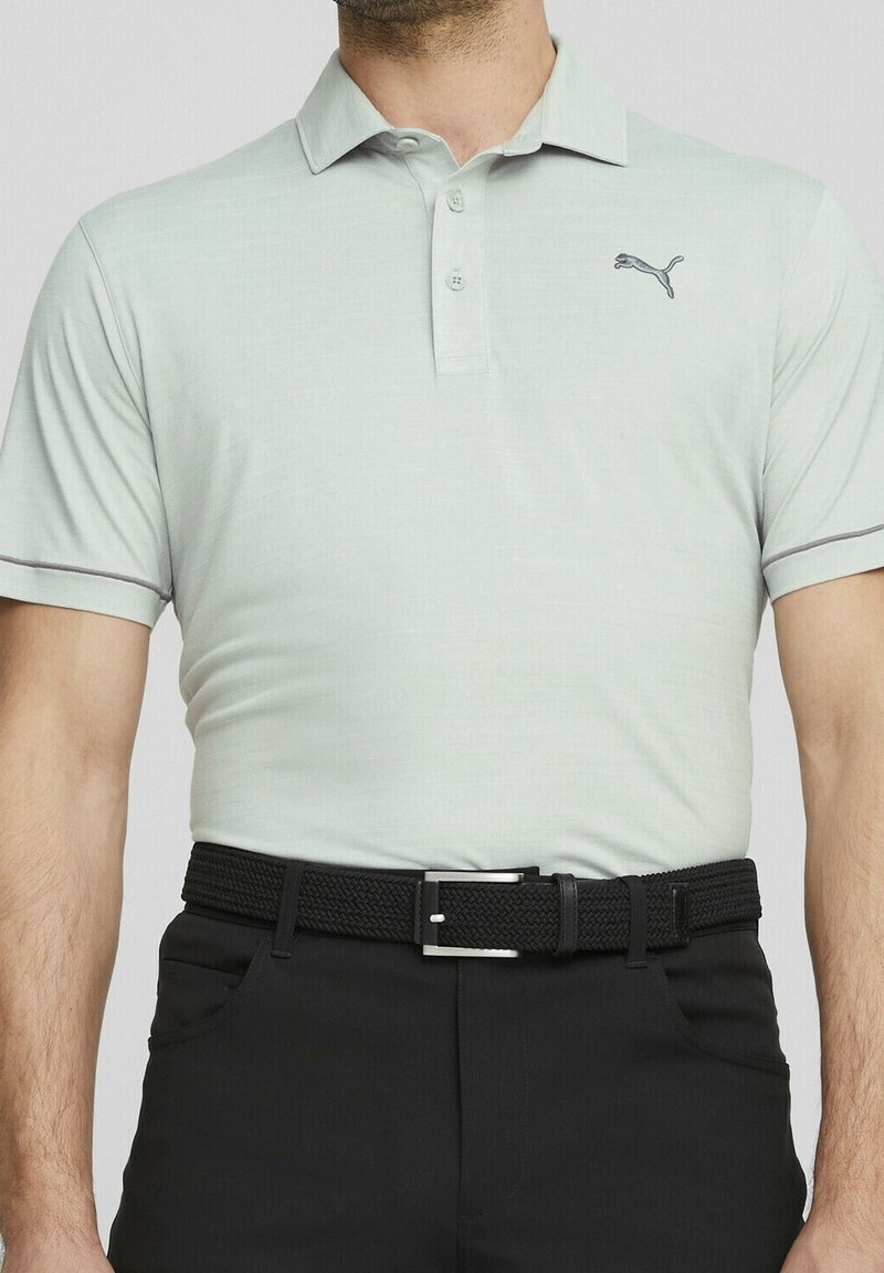 Polo vert clair en tissu texturé, avec un col, trois boutons, et un petit logo Puma bleu marine sur la poitrine gauche. Associé à un pantalon noir et une ceinture tissée noire.