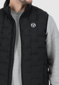 Černá prošívaná vesta se zipovým zapínáním, prošívanou texturou a bílým kruhovým logem na hrudi, doplněná šedou mikinou pod ní.