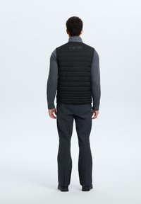 Chaleco de plumas negro con costuras horizontales, combinado con una camiseta de manga larga gris y pantalones oscuros, mostrado desde atrás. Textura suave, diseño elegante.