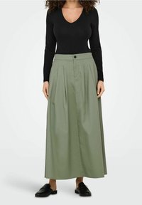 Jupe maxi plissée verte en tissu lisse, avec une fermeture à boutons sur le devant et une ceinture, assortie à un haut noir à manches longues.