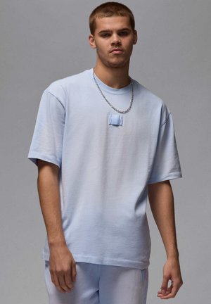 T-shirt oversize azzurro chiaro con colletto girocollo, maniche corte e un dettaglio a toppa sul petto. Abbinata a pantaloni azzurro chiaro. Collana in catena argentata.
